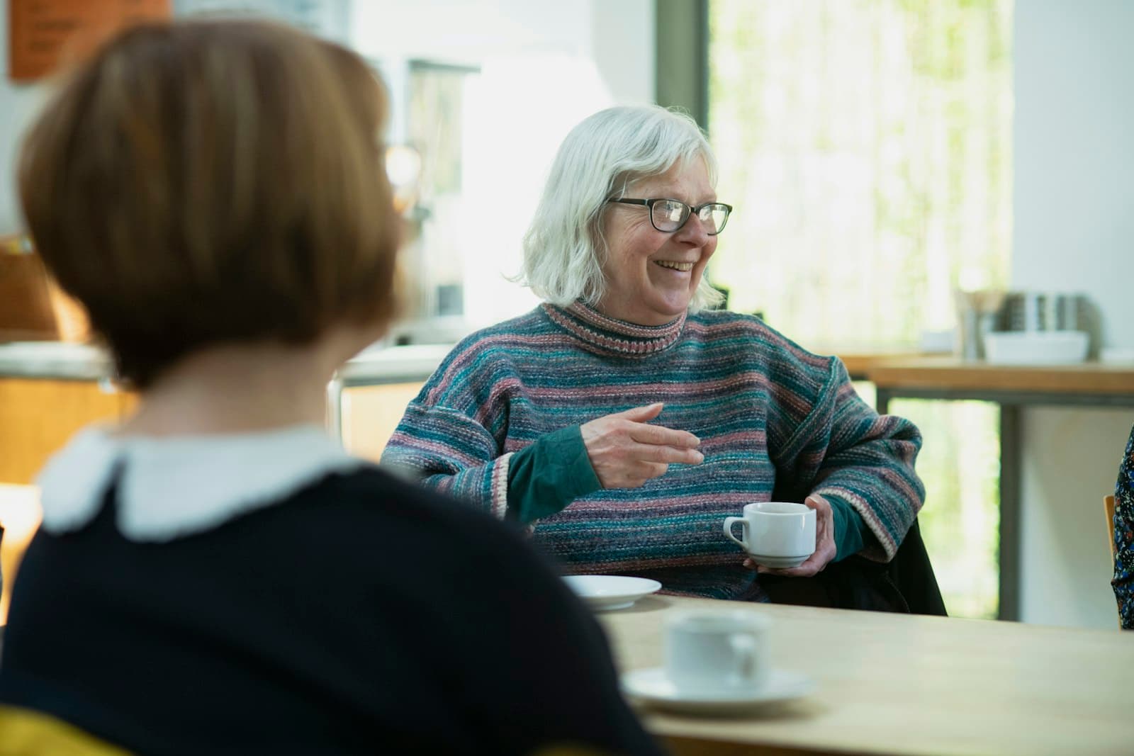 A woman with white hair laughs over a cup of coffee at her kitchen table.