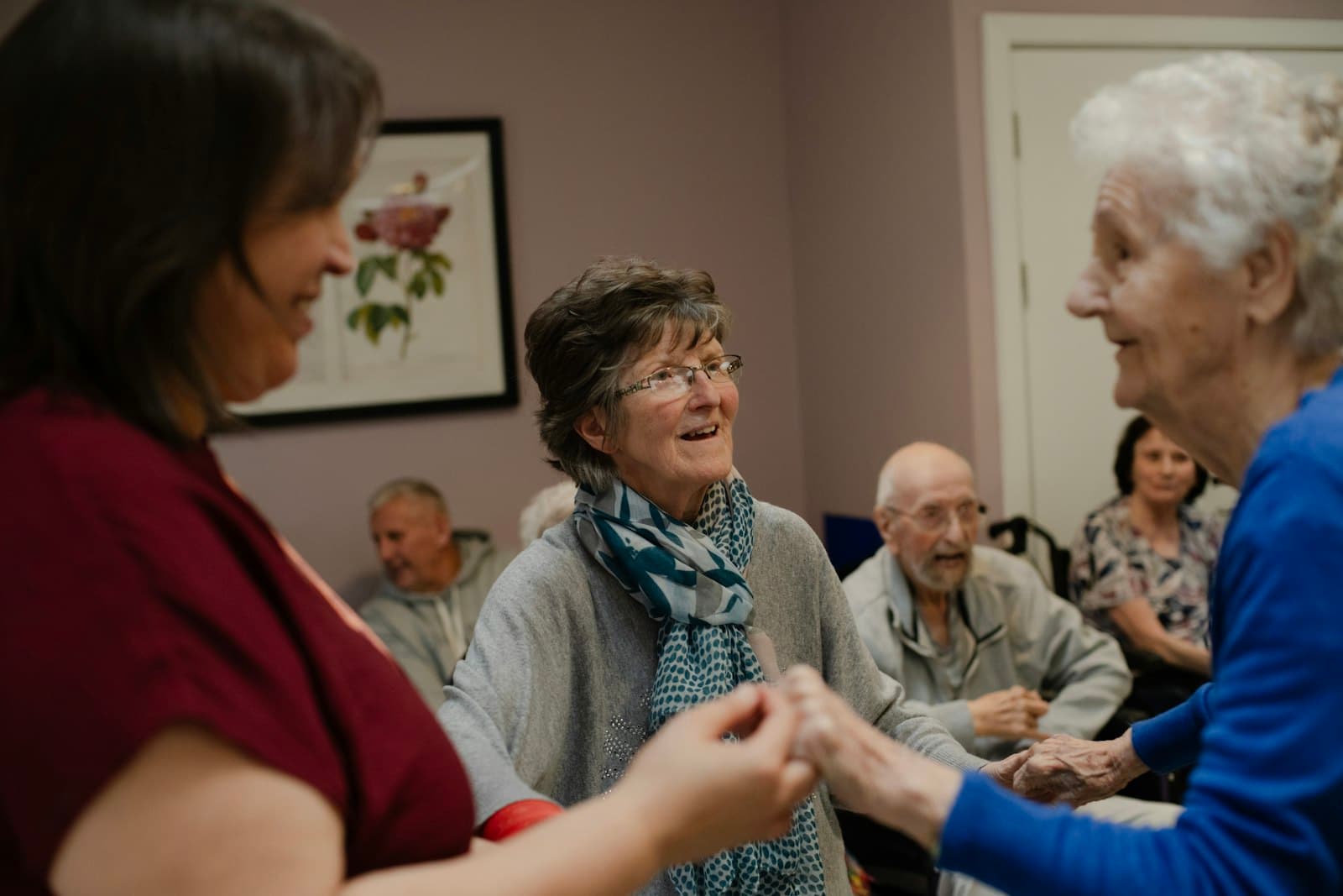 A caregiver holds hands with two women in a bright community room.