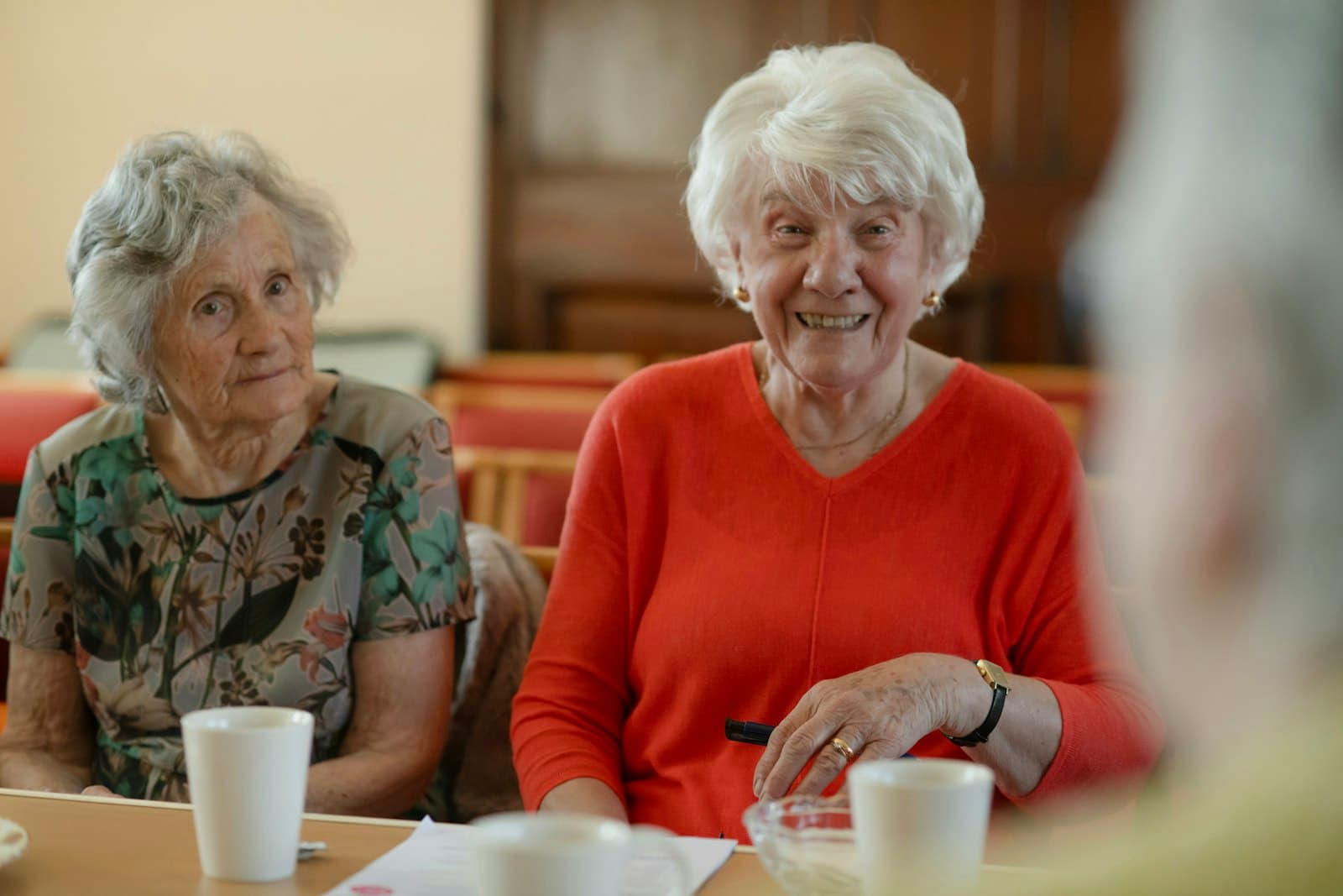 Two older women sitting side by side at a table, one grinning at the camera.