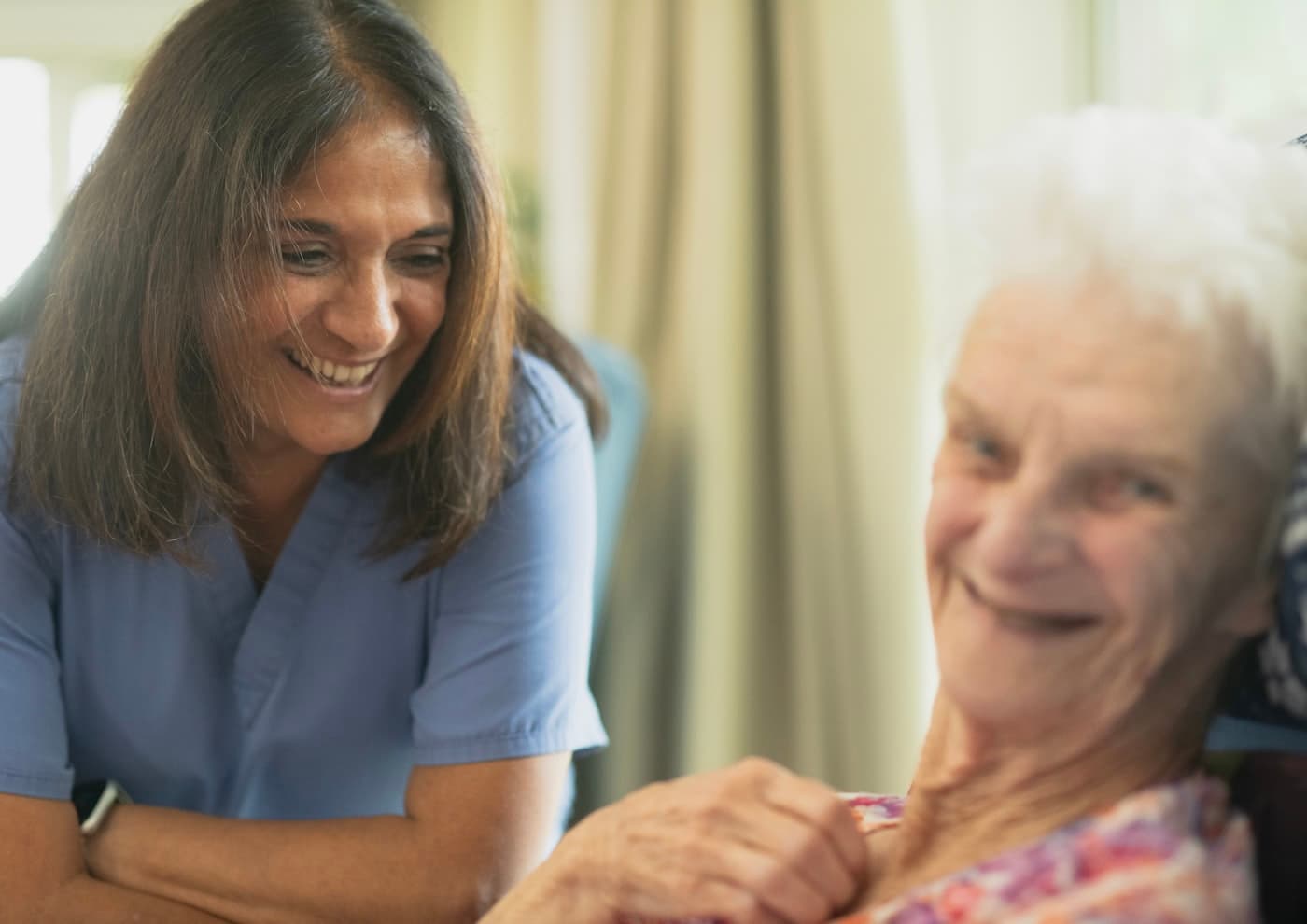 A caregiver in blue scrubs leans in, laughing with an older woman in her armchair.