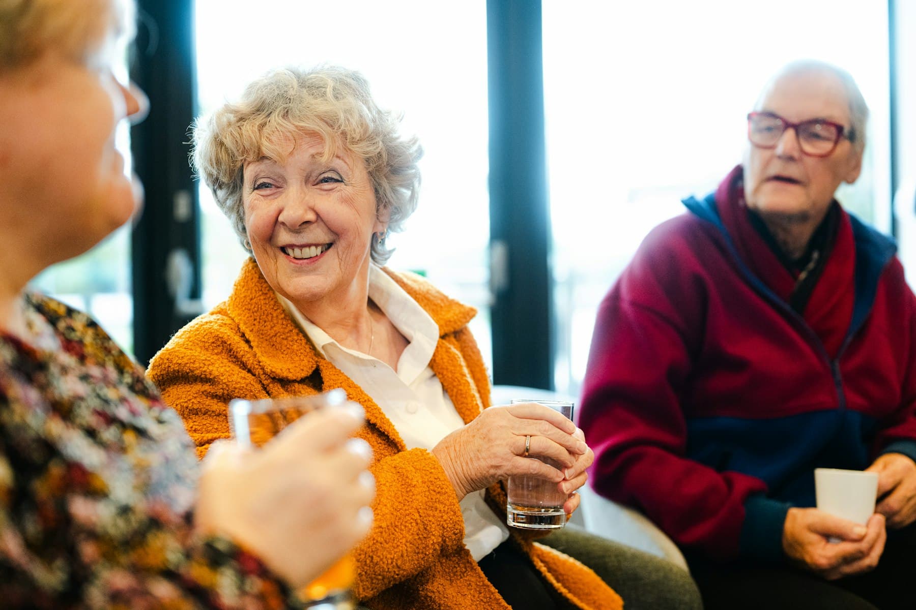 Three older friends sharing drinks and a laugh by a bright window.