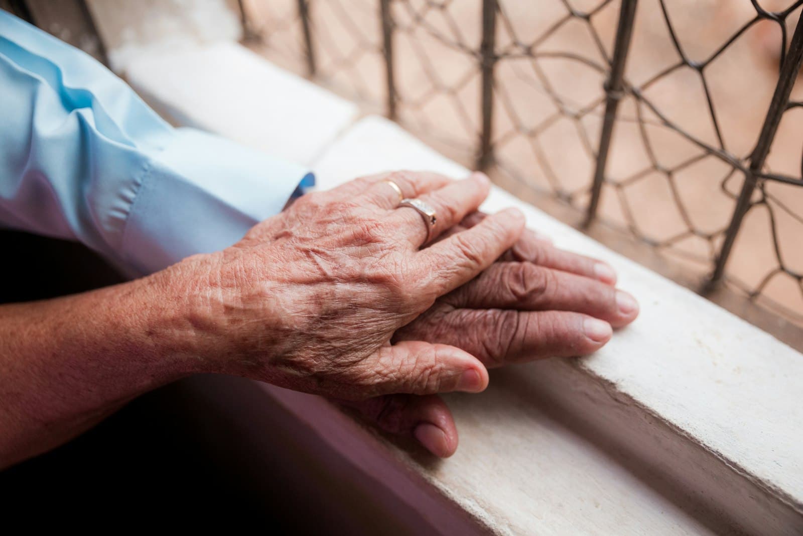An older woman's hands resting gently one over the other on a windowsill.