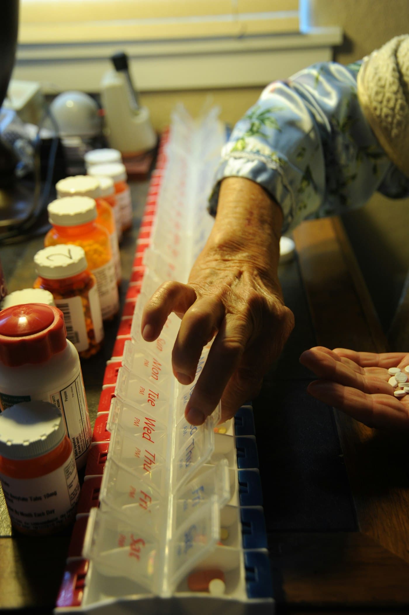 An older woman's hand sorting pills into a weekly organizer beside her prescription bottles.